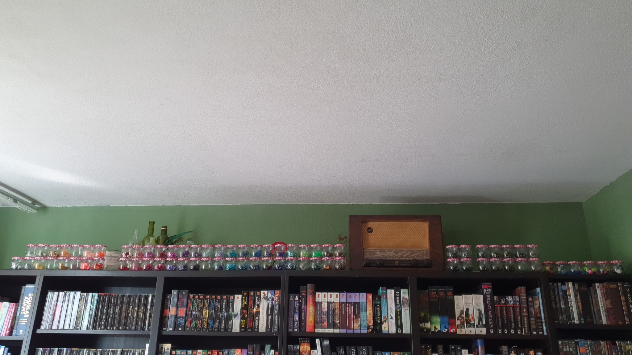 a photo of the top of a row of bookshelves. The top is filled with stacked jars filled with dice. There is also an old radio, and some decorative green bottles.