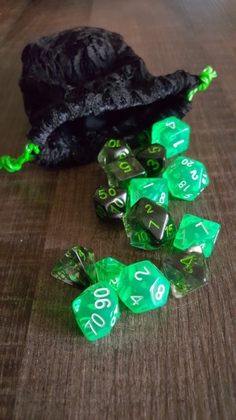 a black lace bag is laying on a wooden table with two sets of dice spilling out. One set is smokey black with bright green numbers. The other is neon green with white numbers