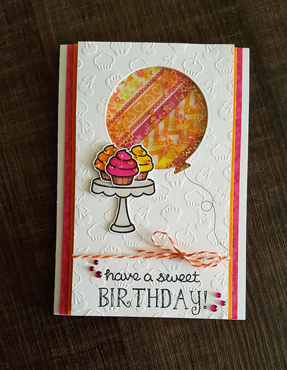 A bright card in orange and pink. A stack of cupcakes sit at the bottom, while at the top a balloon shape is punched out. In the balloon sit glitter and sequins, which move when shaken. The text on the bottom reads Have a Sweet Birthday.