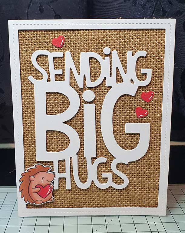 a burlap card with a white border. In the center is a big white text that reads: Sending Big Hugs. In the bottom left corner is a hedgehog holding a heart.