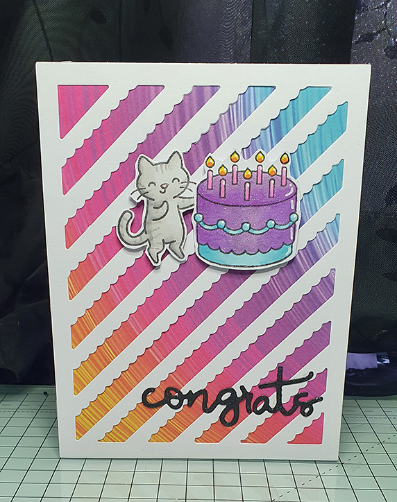 A white card with a striped background in bright colours. A grey cat is holding a bright purple and blue cake. On the bottom is a die-cut of the text "congrats" in black glitter.