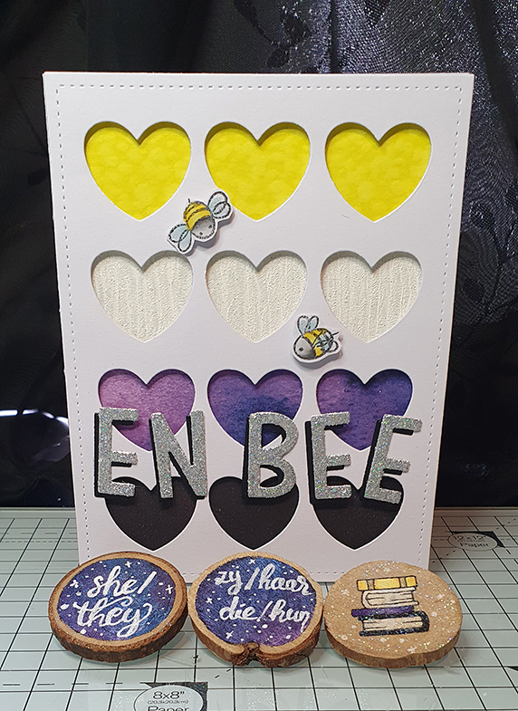 a card with a background of cut out hearts. Behind the hearts are the stripes of the non-binary pride flag. Two little stamped bees fly around. Die-but letters spell out Enbee. In front of the card lay wooden brooches, two with pronouns, and one with a stack of books in the enby colours.