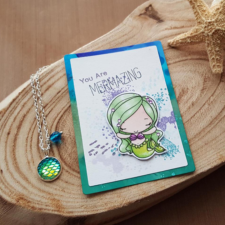 A small Art Trading Card laying on a wooden disk. The background is greens and blues. On the card is a mermaid with her arm wide and her eyes closed. Her hair and tail are green, she is wairing a purple shell bra. Next to her is text saying You Are Mermazing.