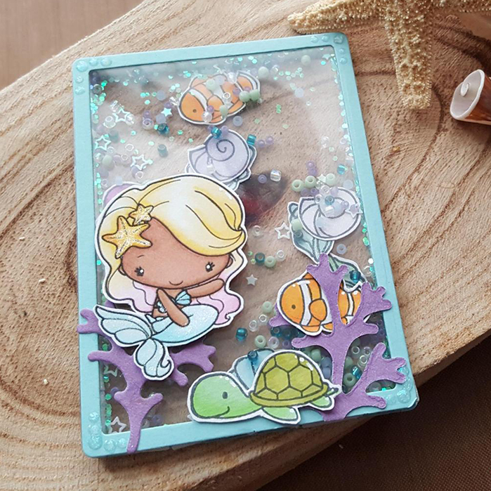 A small transparent art trading card, with a stamp of a mermaid. Inside the card are beads, sequins and small stamped shells and fish, which move when shaken.