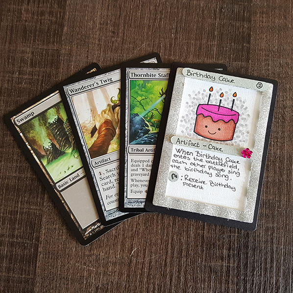 A small birthday card made in the same style as a Magic the Gathering card. The top has a pink birtday cake in it, the text says "artifact-cake. When Birthday Cake enters the battlefield, each other player sings the birthday song. Tap: Receive birthday present"