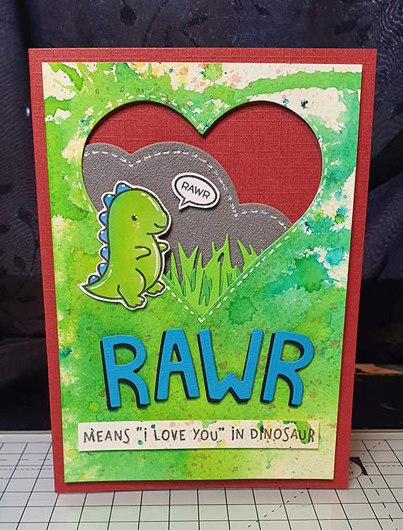 A red card with a watercolour green backgound on top. A heart is cut out, making a window through which you see grass and rocks. A small green dinosaur with blue spikes sits next to it. Below it reads RAWR means "I love you" in dinosaur!