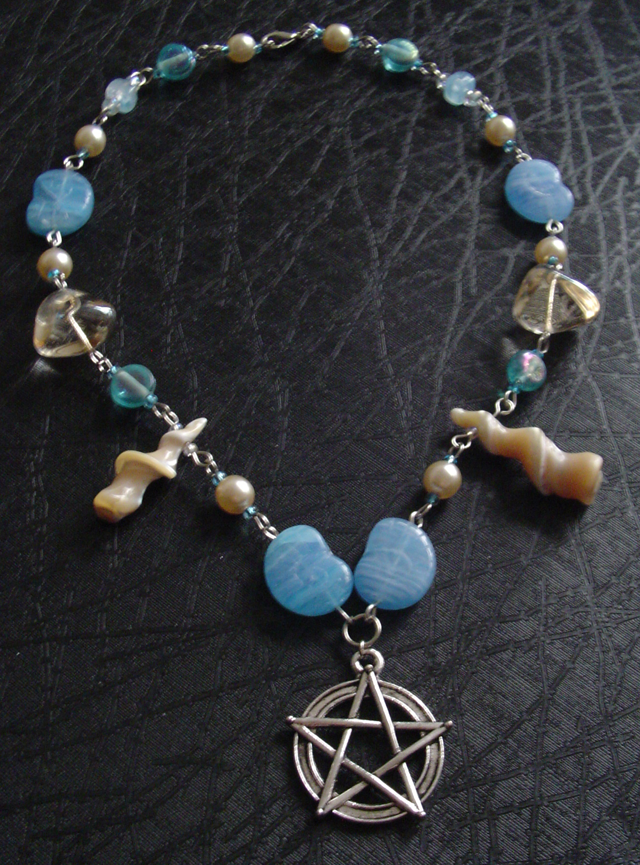 A beaded necklace with blue and shell coloured beads in various shapes and sizes. The centre pendant is a pentacle. Two small twisted shells hang on both sides.