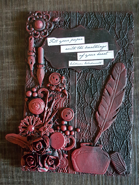 a black and red card made with mixed media. On the left there are a lot of buttons, quill nibs, and other metal parts which have all been spraypainted black, and then stained red. On the right sits an inkwell and quill. In the center is a text on white paper that reads: Fill your paper with the breathings of your heart. ― William Wordsworth