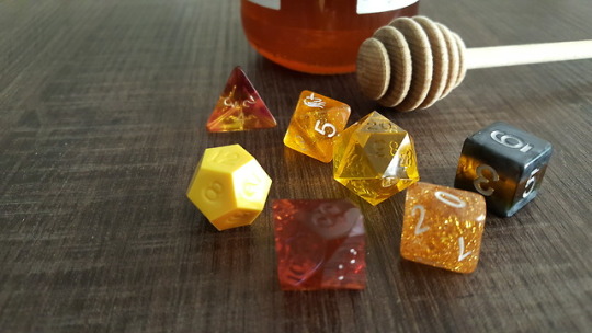 a set of mixmatched dice laying on a brown surface. A pot of honey sits in the background. A honey dipper is laying next to the dice. The dice are in order: sharp edge clear yellow, sharp edge opaque yellow, sharp edge clear brown, glitter gold, glitter gold, black and yellow striped, clear brown and yellow.