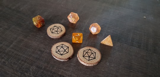 three wooden tokens laying on a wooden table together with some yellow dice. The tokens feature a drawn d20 in black, with golden rays radiating out