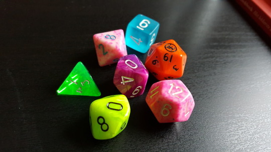 a set of mixmatched dice laying on a black surface. The dice are in order: bright orange, neon yellow, neon purple, speckled pink, speckled pink, frosted blue, and neon green.