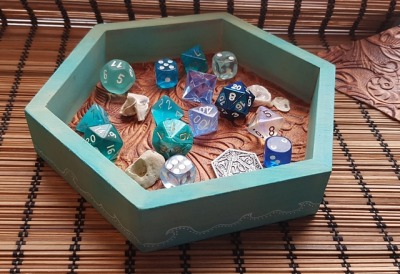 a hexagon shaped dice box on a wooden surfave. Inside is a set of mismatched dice is various shades of seablues, some shells, and a d20 coin
