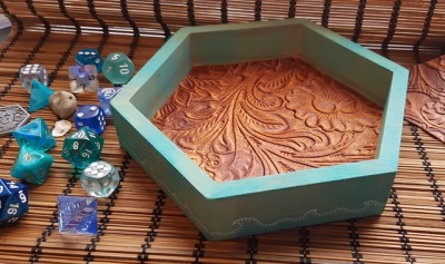 a hexagon dice tray in greens and blues with waves on the side. The inside is covered in brown leather stamped with swirls. There are blue dice laying next to the dicebox